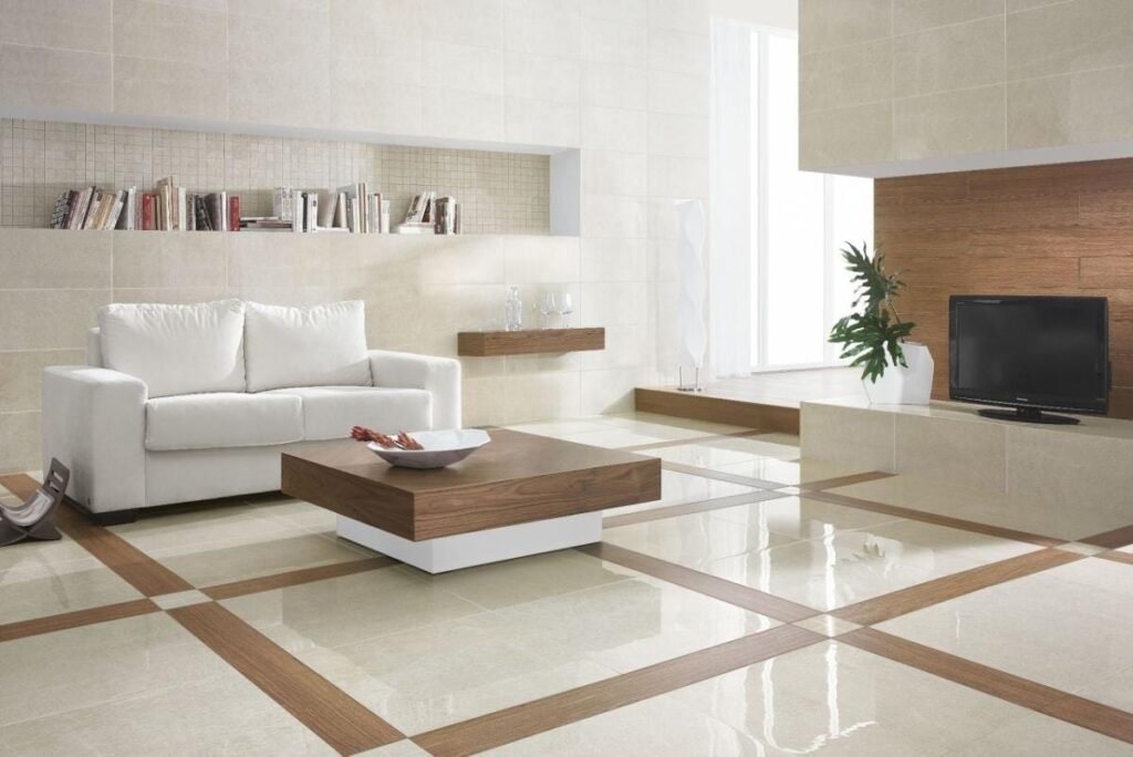 Modern living room with a white sofa, wooden coffee table, and TV. Beige tiled walls and floors, books on a shelf, minimalist decor.